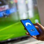 Osoba drži telefon dok u pozadini gleda fudbalsku utakmicu na televizoru