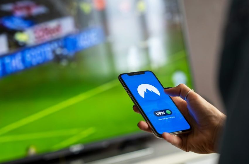 Osoba drži telefon dok u pozadini gleda fudbalsku utakmicu na televizoru