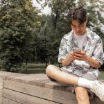 Muškarac u košulji i bermudama sedi na klupi i gleda u mobilni