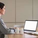 Žena sedi za stolom pored laptopa i dve bele bočice sa sveskom i čašom vode