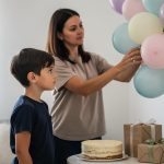 Žena kači pastelne balone na zid dok dečak posmatra pored torte i poklona