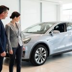 Muškarac i žena u odelima stoje pored srebrnog automobila u salonu